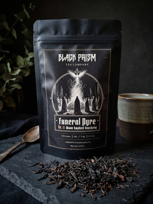 A black stand-up pouch of Black Prism Tea Company 'funeral Dyre' No. 11 Macbethoed Black Tea is displayed on a dark slate surface with loose tea scattered in front and a ceramic cup to the side.