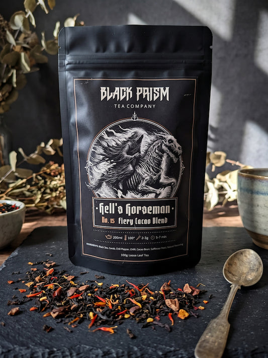 A black stand-up pouch of Black Prism Tea Company 'Hell's Horseman' No. 15 Fiery Loco Blend tea, displayed on a dark slate surface with loose tea leaves and a spoon.